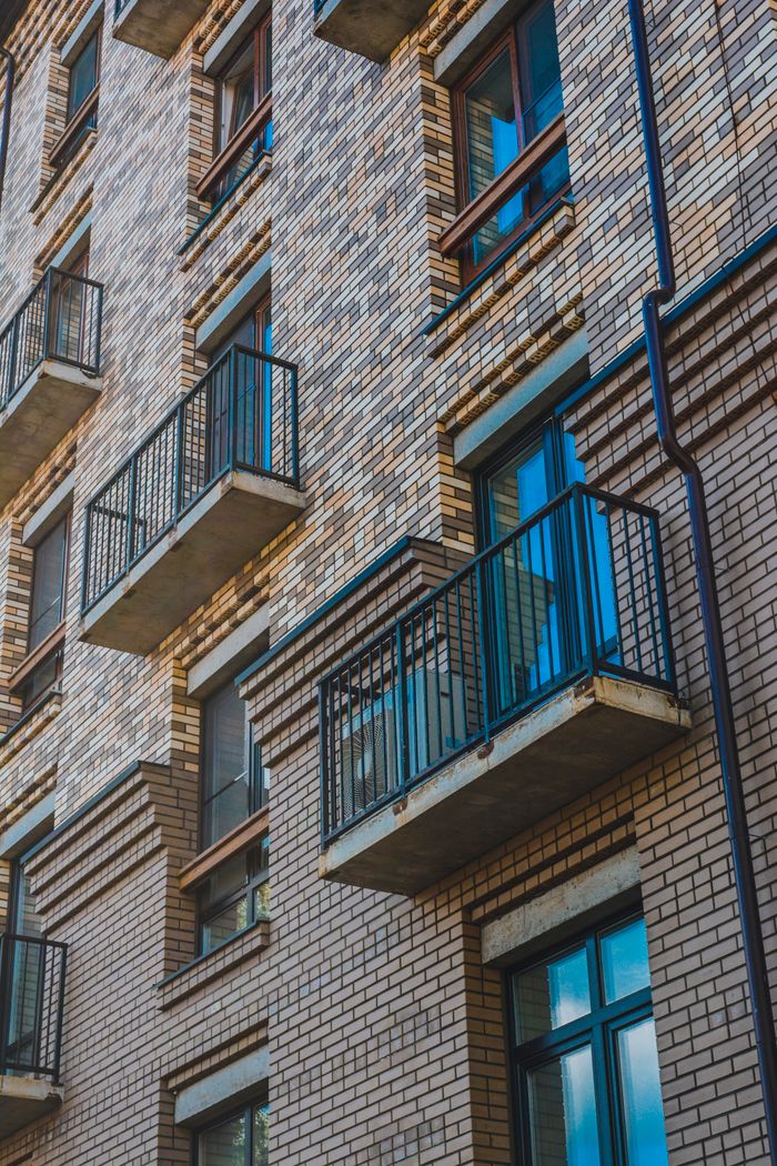 contact-img Low-angle view of a brick apartment building with balconies and metal railings, showcasing a modern urban design.